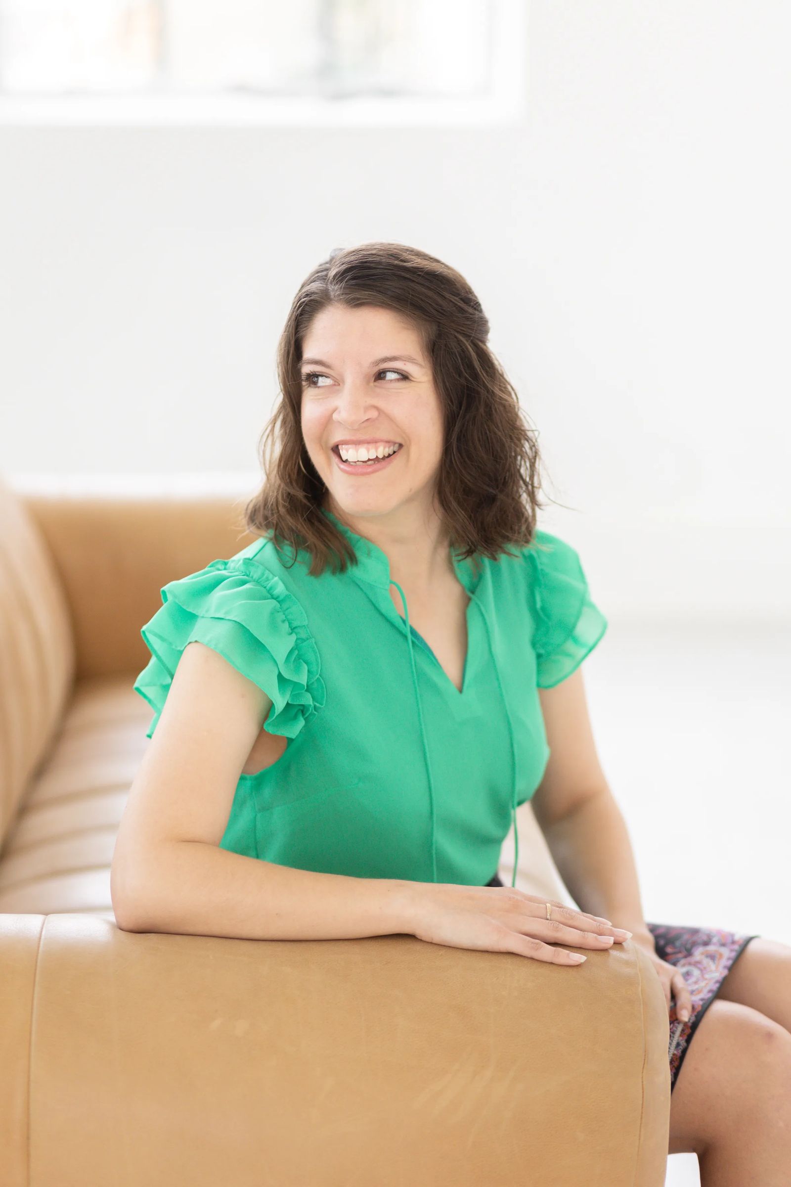 Smiling female looking to the side while sitting on a light brown couch. She is wearing a green top, has short brown hair.